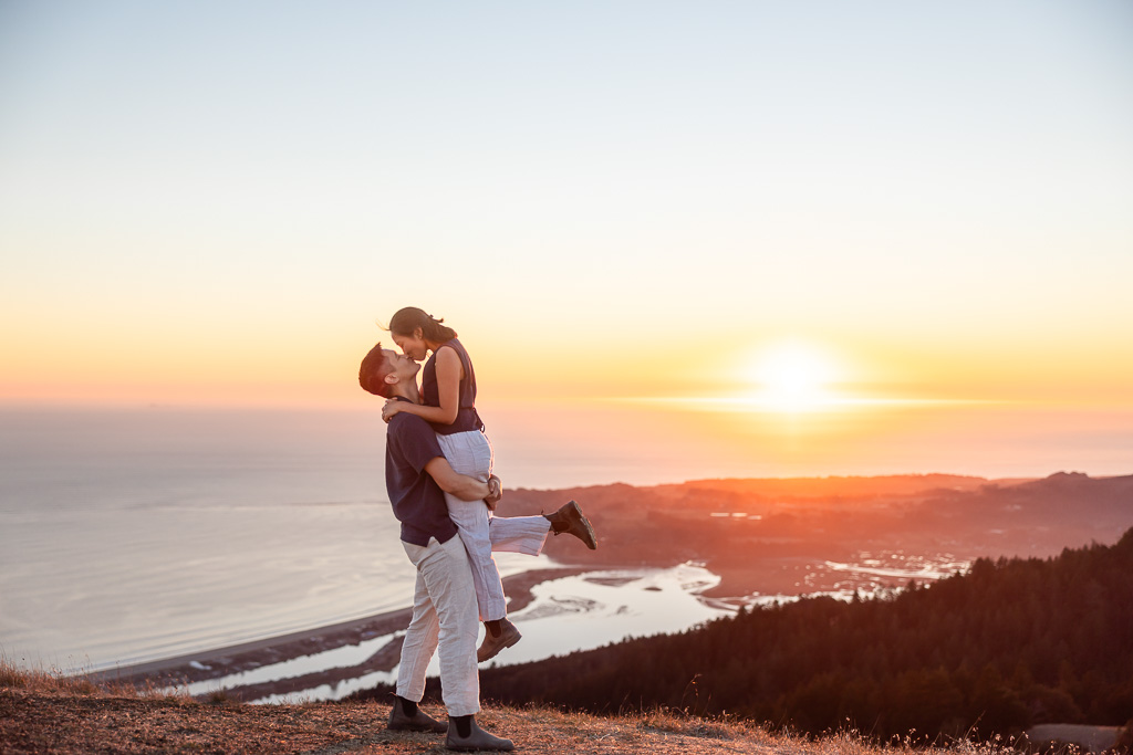 A woman wraps her arms around her partner as he lifts her up beside the sunset view over the coast.