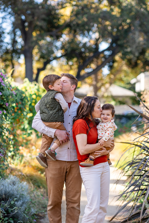 Fall family photos at Elizabeth F Gamble Garden show both parents sharing a kiss with their young children along a leafy garden path.