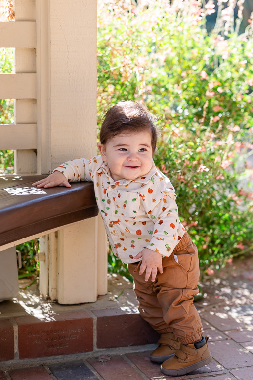 A smiling baby stands beside a wooden garden bench, framed by green foliage and dappled autumn light.