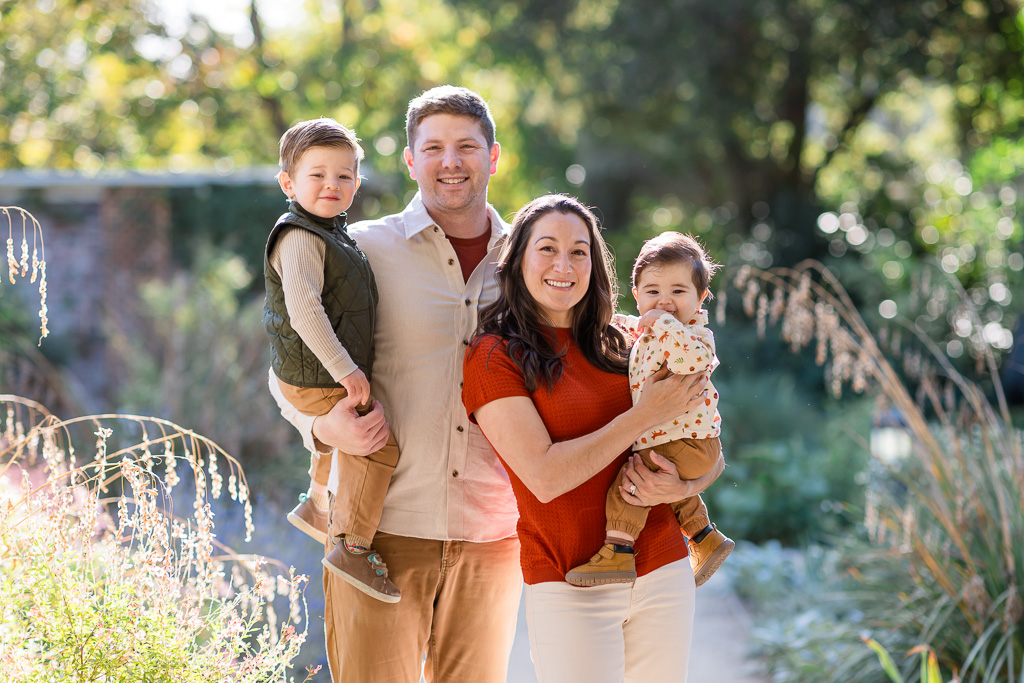 Holiday family photos capture two parents holding their children for a sunlit portrait in the garden.