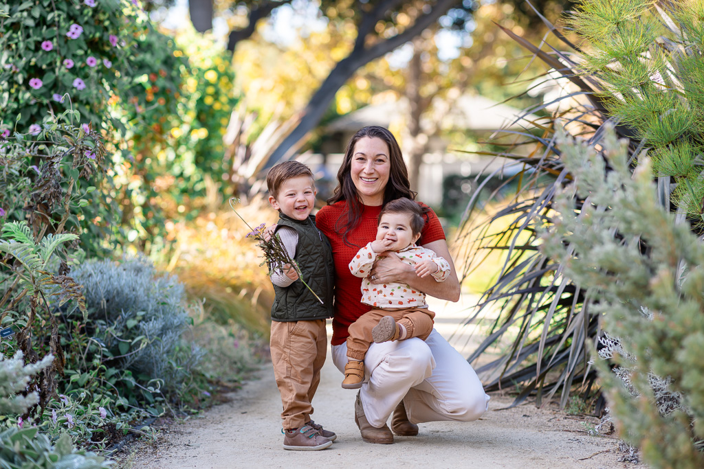 A mother sits on a garden path holding her baby while her older boy stands beside her.