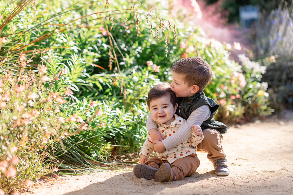 An older sibling leans in to kiss the baby’s cheek on a sunny garden path.