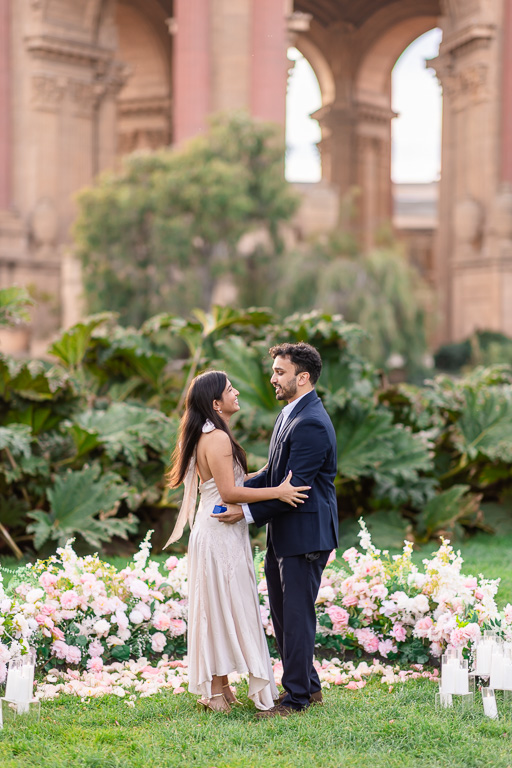 A couple faces each other among pink and white flowers beneath the arches of the Palace of Fine Arts.