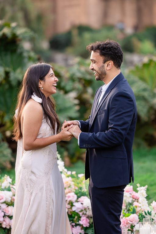 The couple holds hands and laughs together in a candid moment while surrounded by pastel proposal flowers.