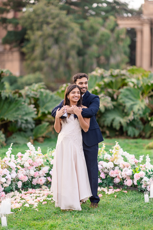 A man embraces his smiling partner from behind as they pose among flowers for their engagement portraits.