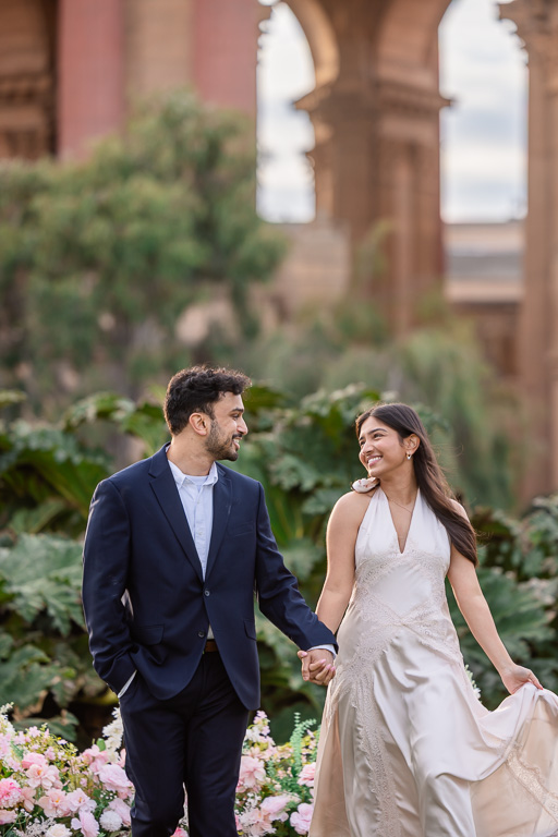Golden hour engagement photo of couple walking hand in hand, smiling at each other as she lifts the skirt of her dress.