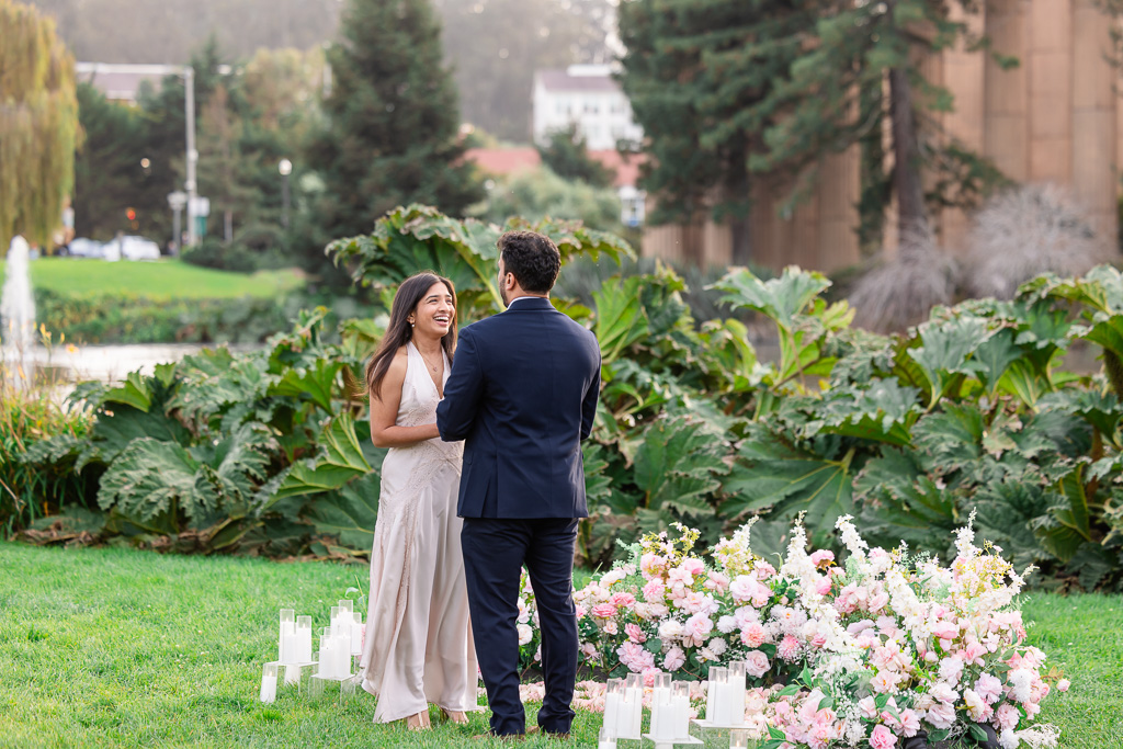 A couple laughs beside pink and white floral proposal decor by Something Special Picnics on the Palace of Fine Arts lawn.