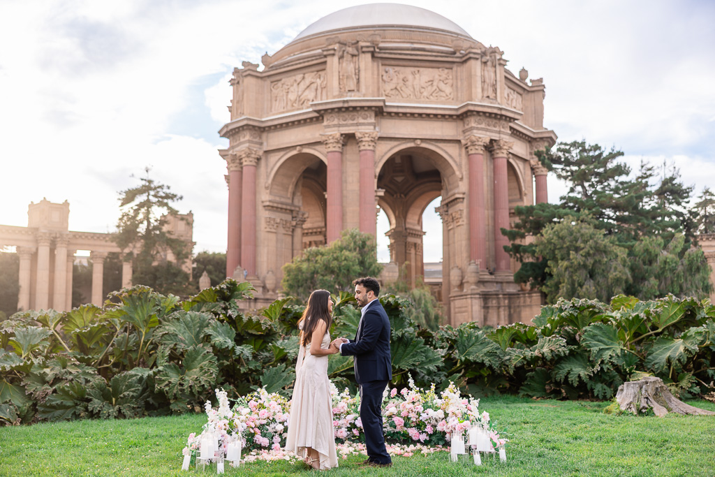 Wide view of a couple holding hands inside a floral proposal setup beneath the Palace of Fine Arts rotunda.