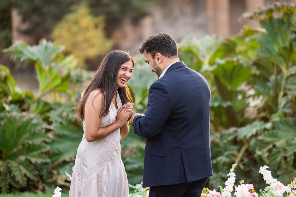 A girl laughs with her hands clasped near her heart as her fiance pull out the ring box before the proposal.