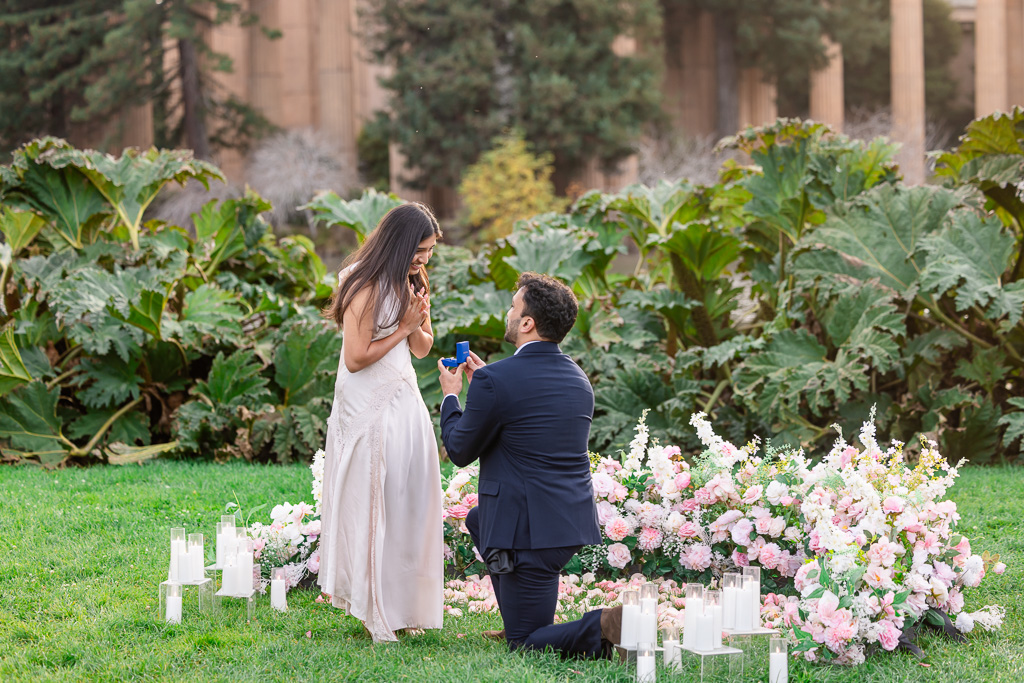 A man kneels with a ring box beside pink and white flowers by Something Special Twins as his partner clasps her hands.