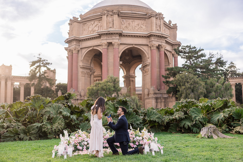 A man proposes on one knee amid flowers and glass candle holders, with the Palace of Fine Arts at golden hour as the backdrop.