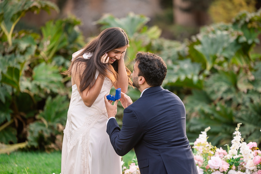 A delighted woman raises her hands to her face as her kneeling partner presents an engagement ring in a blue box.