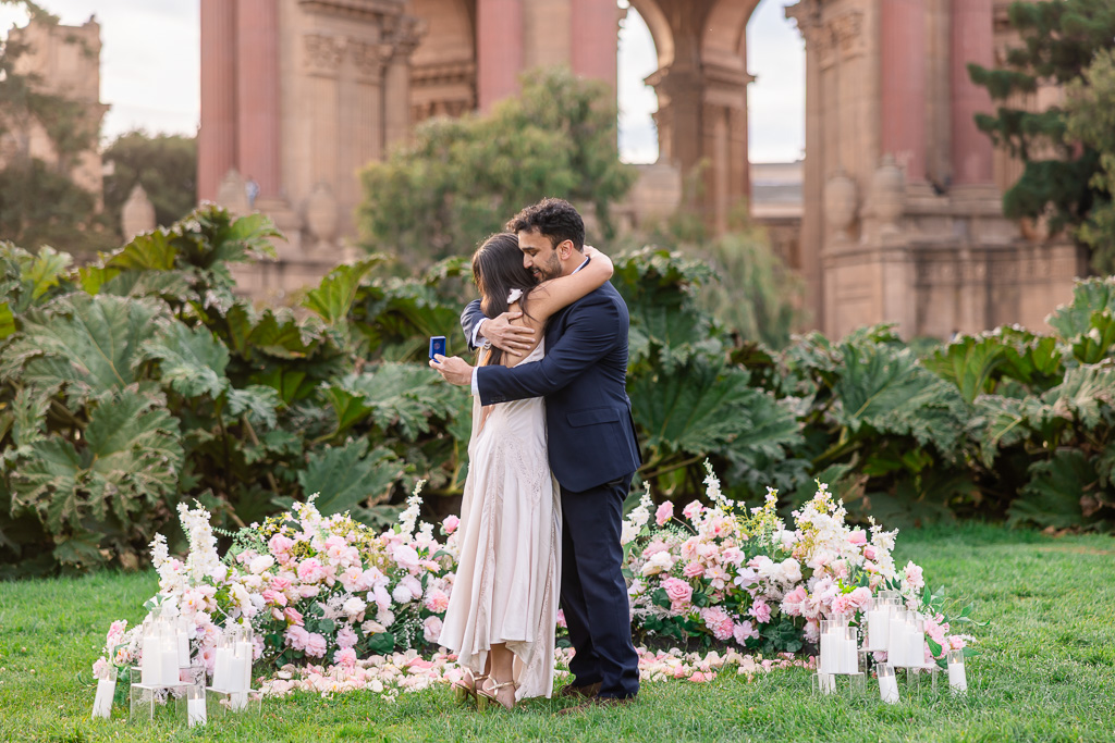 The couple shares a tight embrace after the proposal, surrounded by pastel flowers and glass candle holders.