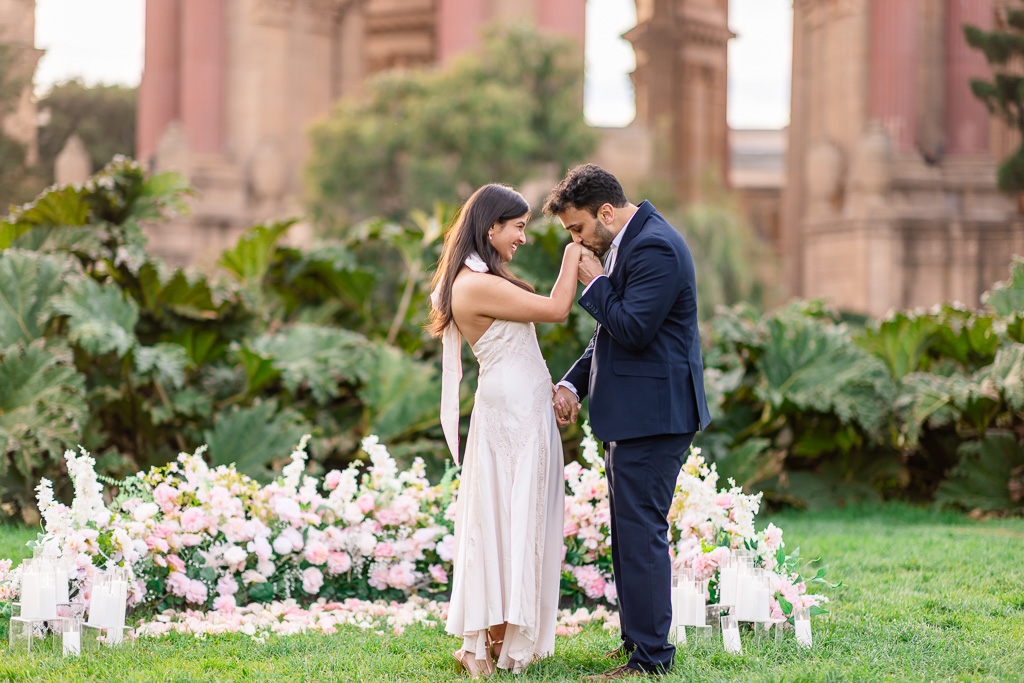 A man kisses his newly engaged partner’s hand as she smiles in front of the Palace of Fine Arts arches.